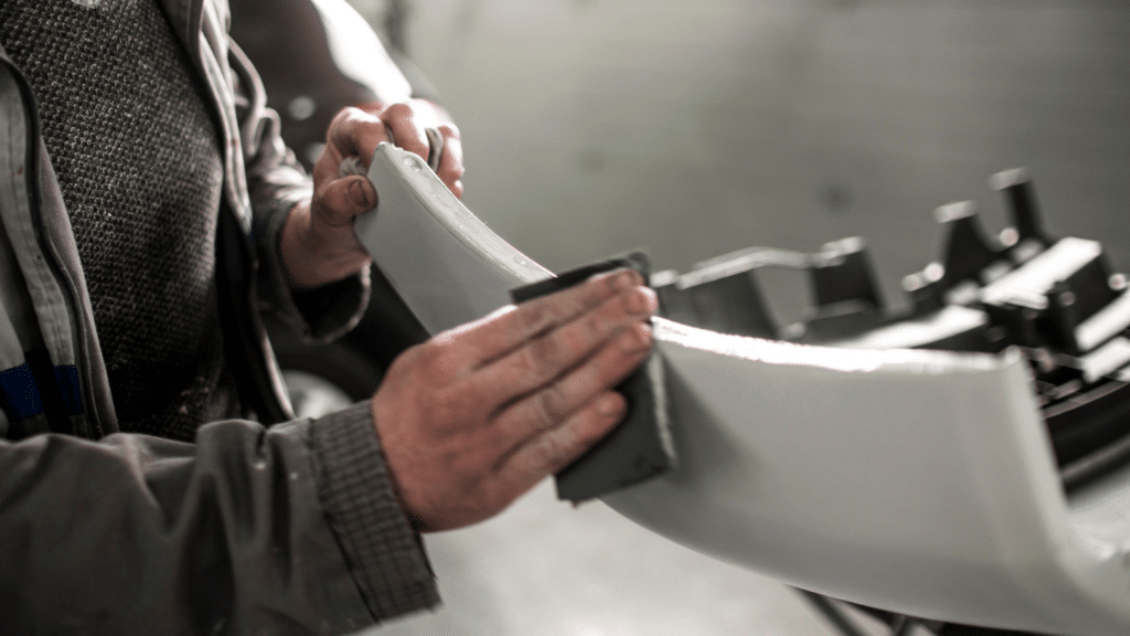 Body repair technician removing excess filling from an accident-damaged car bumper using a sanding paper.